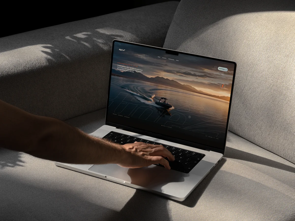 Hand on a laptop on a sofa showing the Tigé wakesurf boats homepage at sunset